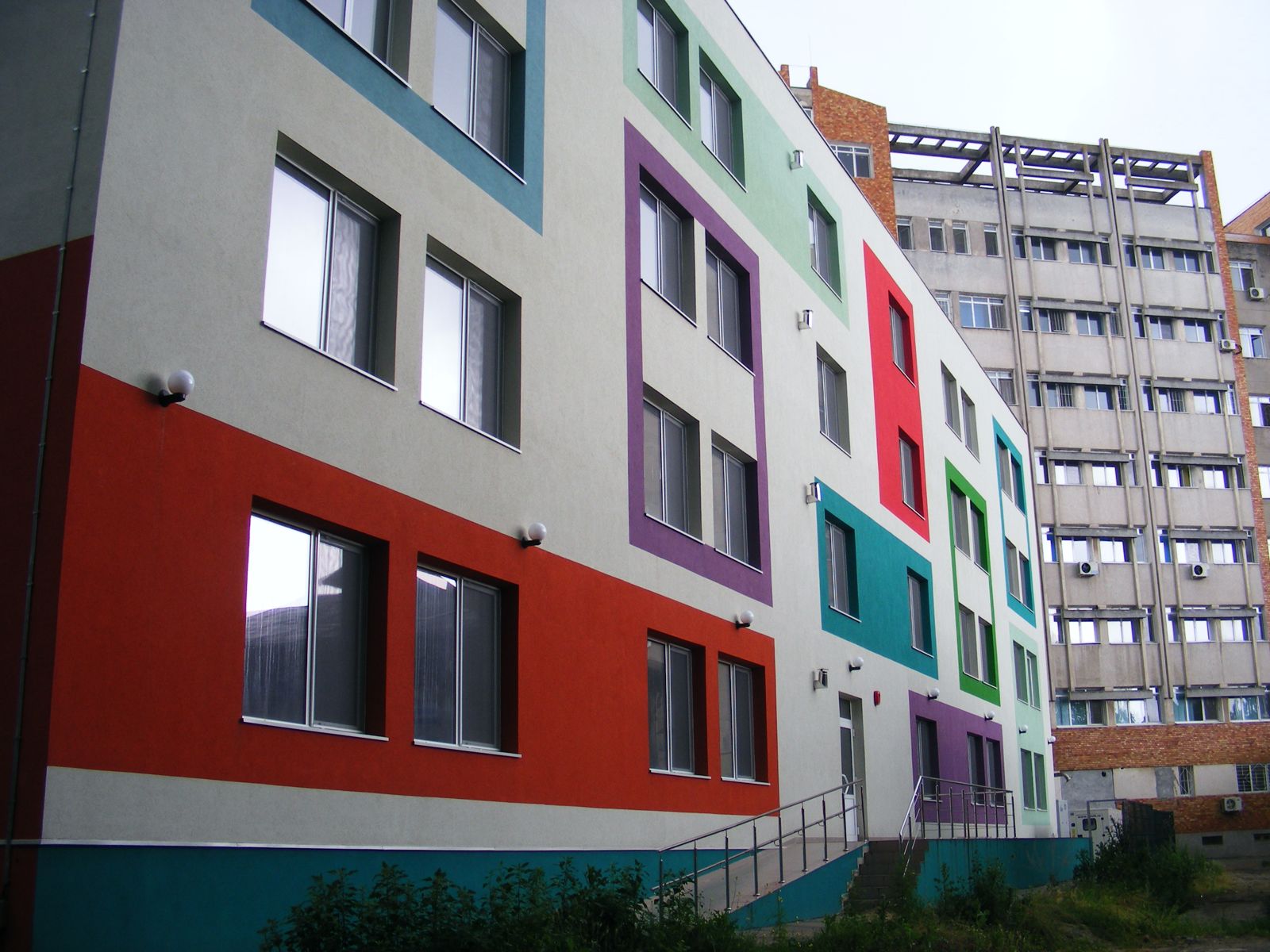 Fațada colorată a unui corp nou de patru niveluri, cu clădirea mai înaltă a corpului existent în fundal